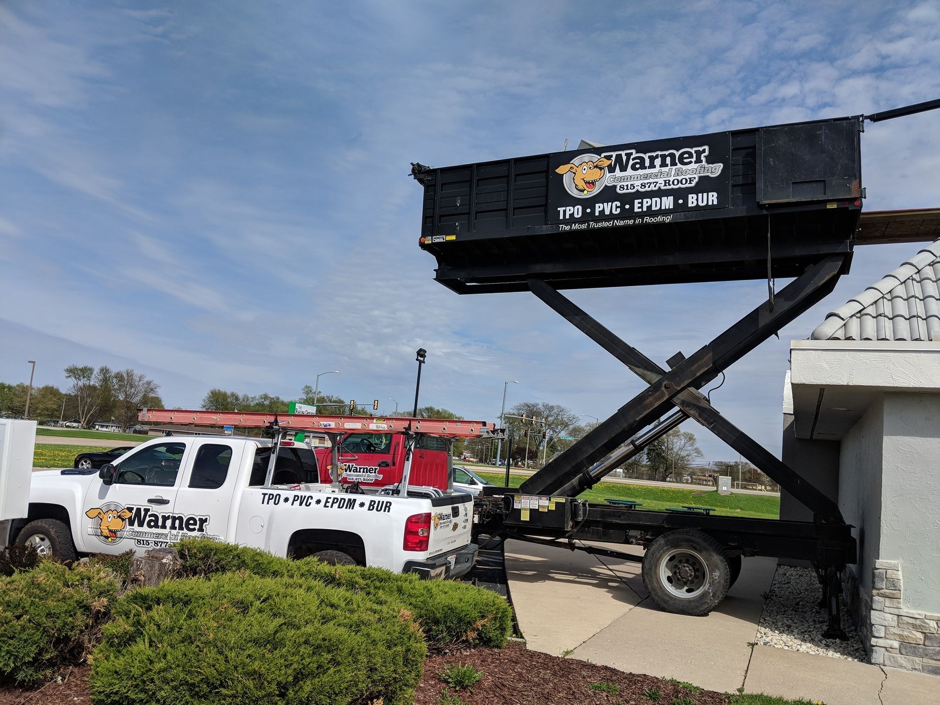 A white pickup truck hauls a black dumpster lift elevated to the roof of a building.