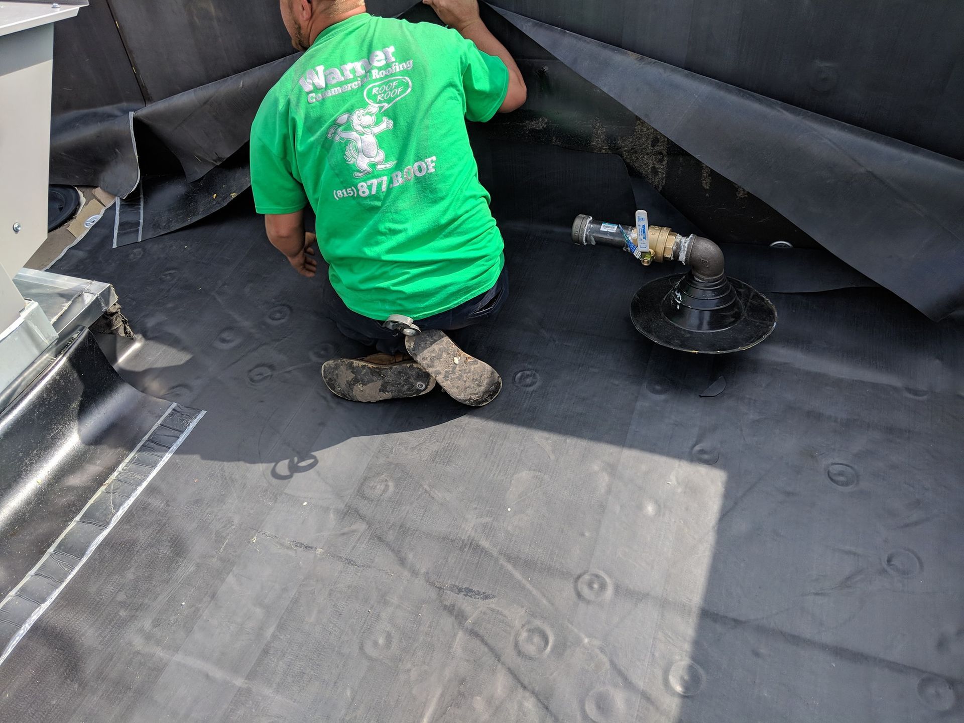 A worker in a green shirt kneels on a black rubber roof surface near a pipe fitting and flashing.