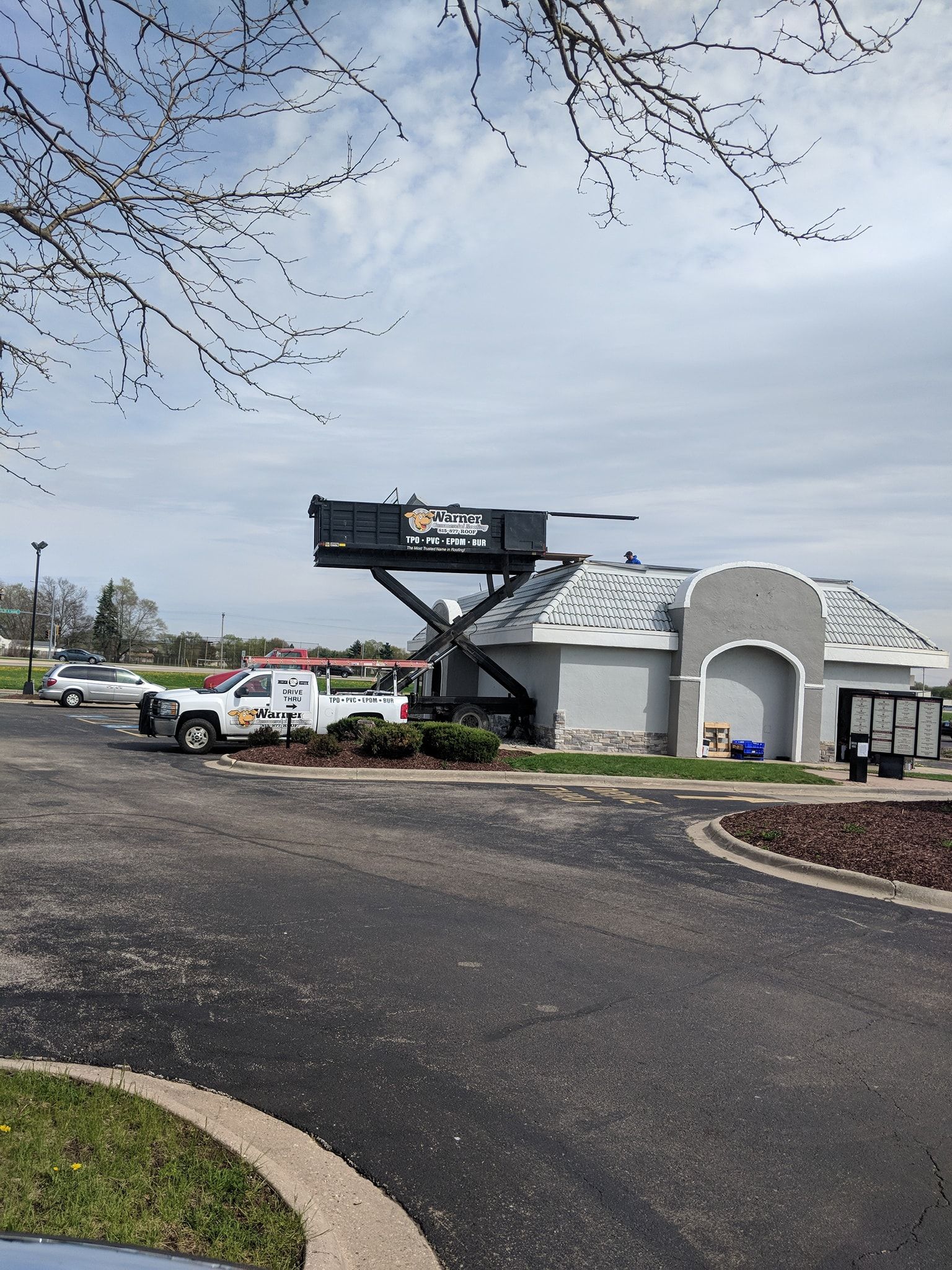 A dump trailer with a raised hydraulic lift bed sits next to a commercial building under a cloudy sky.