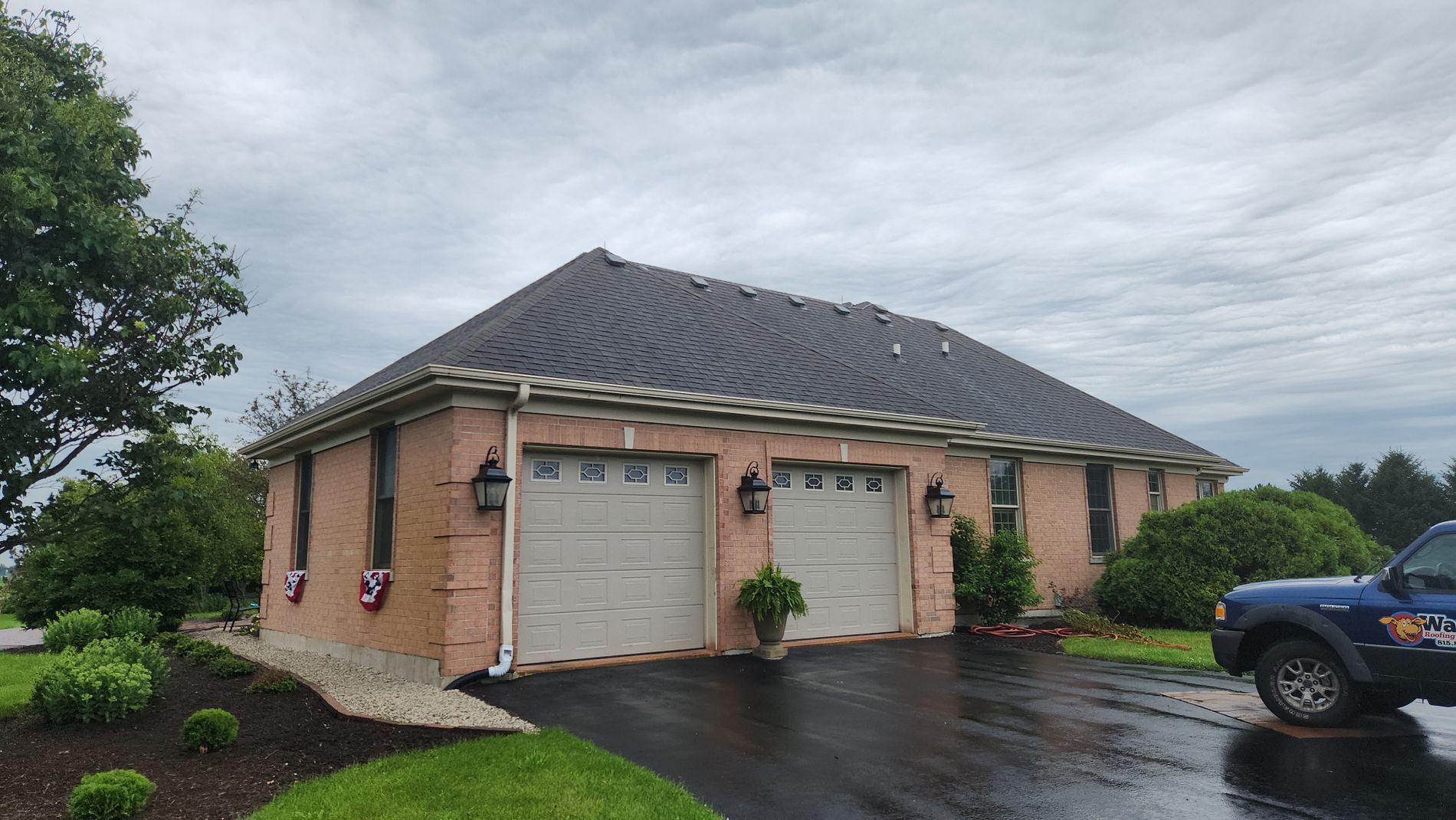 A brick house with a two-car garage, gray roof, and landscaped lawn under a cloudy sky.