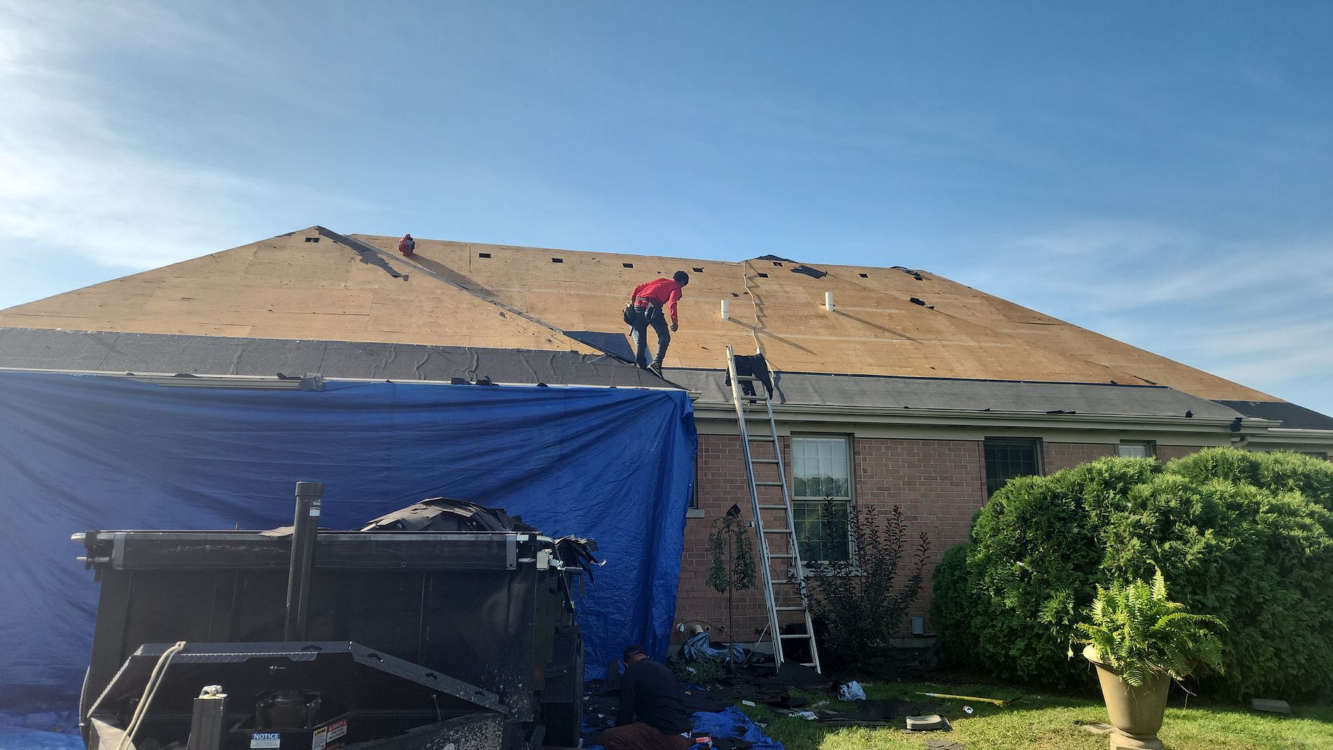 A worker in a red shirt walks on a residential roof under repair, with a ladder and blue tarps nearby.