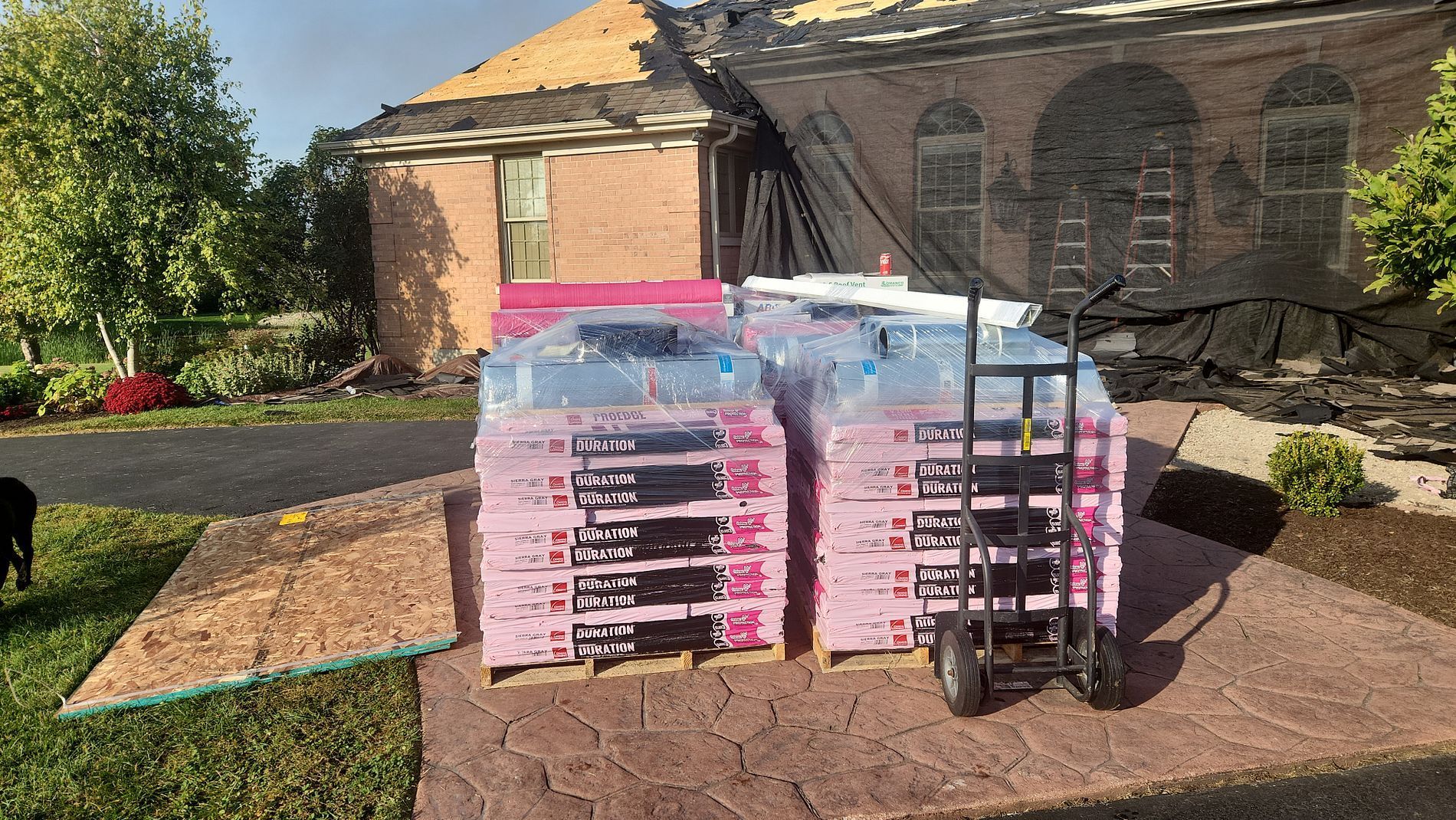 Stacked pink insulation boards on a pallet sit in front of a house undergoing roof repairs on a sunny day.