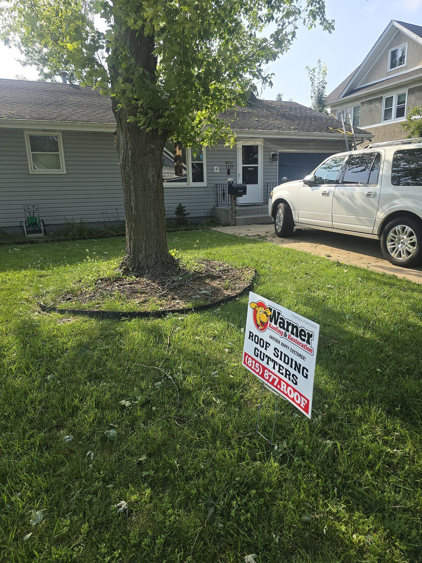 A white SUV is parked on a driveway in front of a gray house with a lawn sign for a local service company.