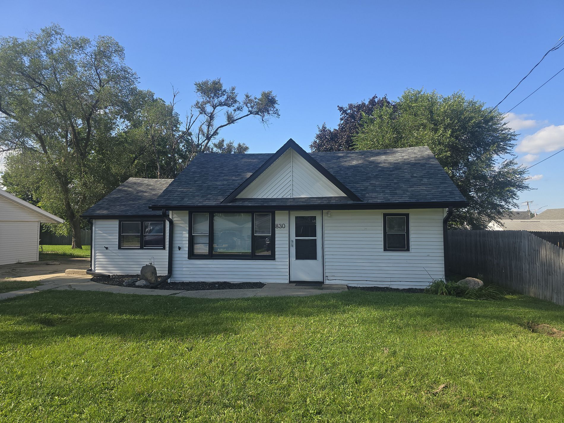 A small, white, one-story house with black trim, a gabled roof, and a front lawn under a clear blue sky.