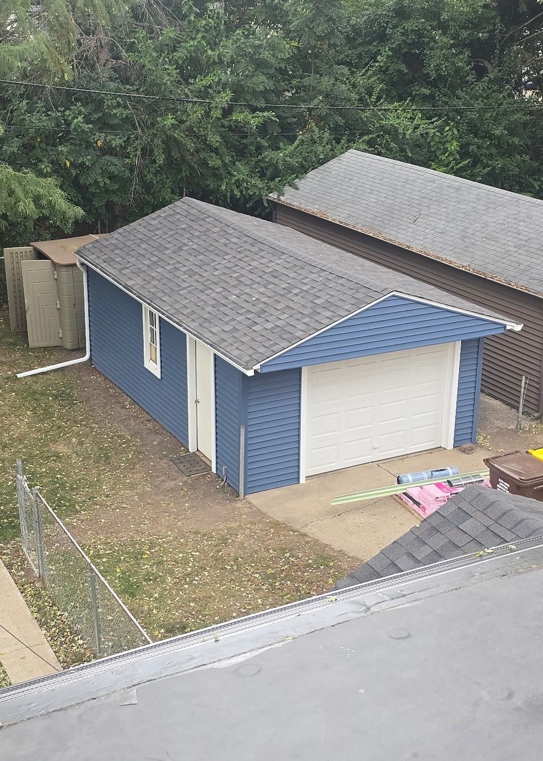 A blue, detached garage with a white door and gray roof sits in a grassy yard next to another garage.