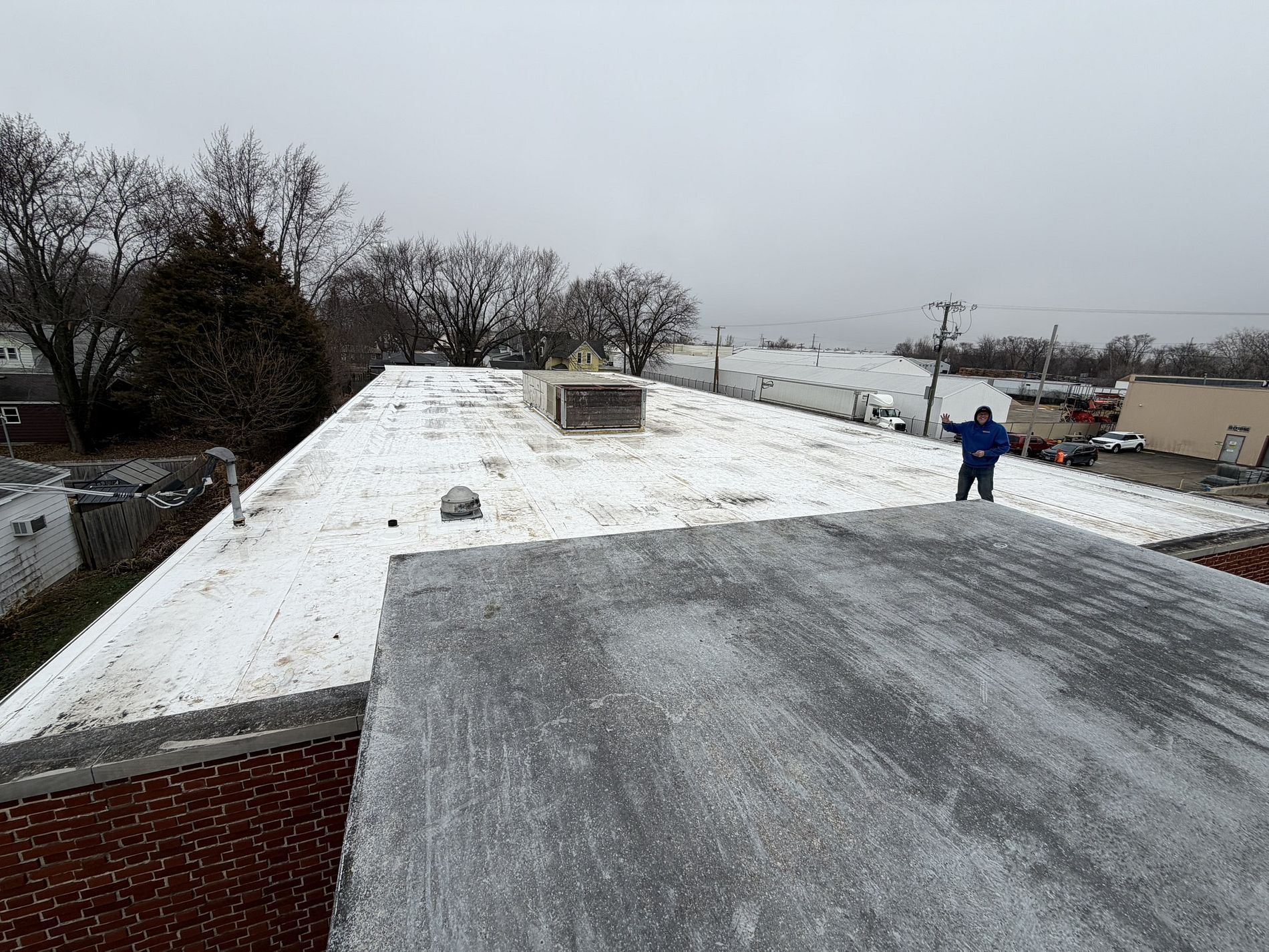A person in a blue jacket walks on a white, weathered flat roof toward a rectangular vent on a cloudy day.