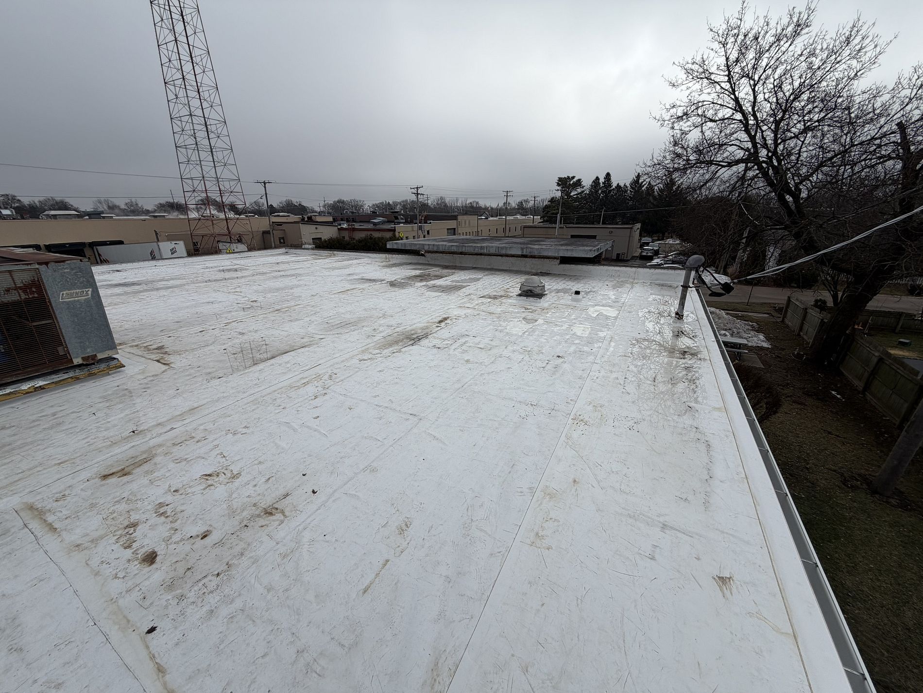 A white TPO flat roof under an overcast sky, with a metal drip edge, nearby utility equipment, and distant trees.