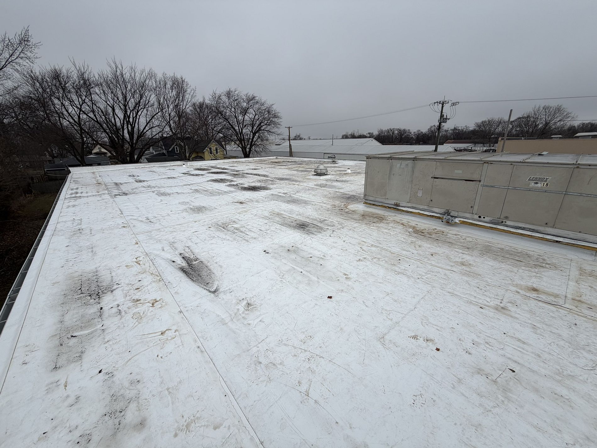 A white flat roof covered in a light layer of snow under a grey sky, with a rooftop HVAC unit visible on the right.