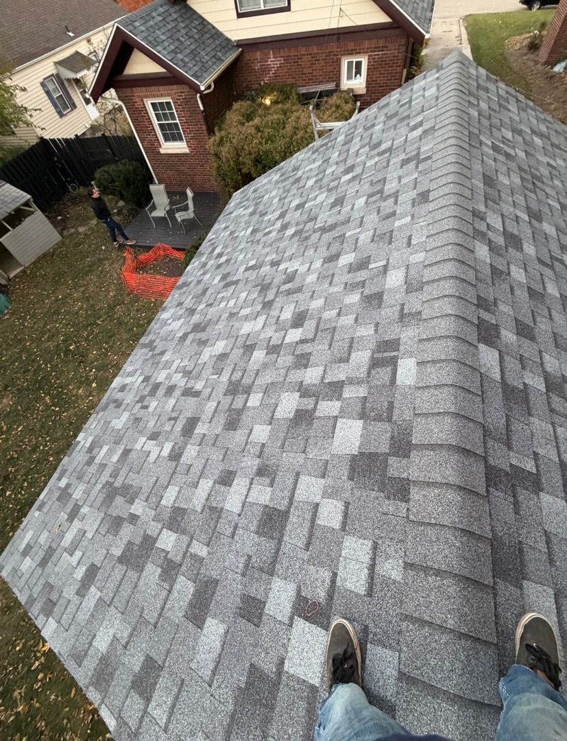 A person stands on a grey shingled roof looking down at a backyard and a neighboring house.