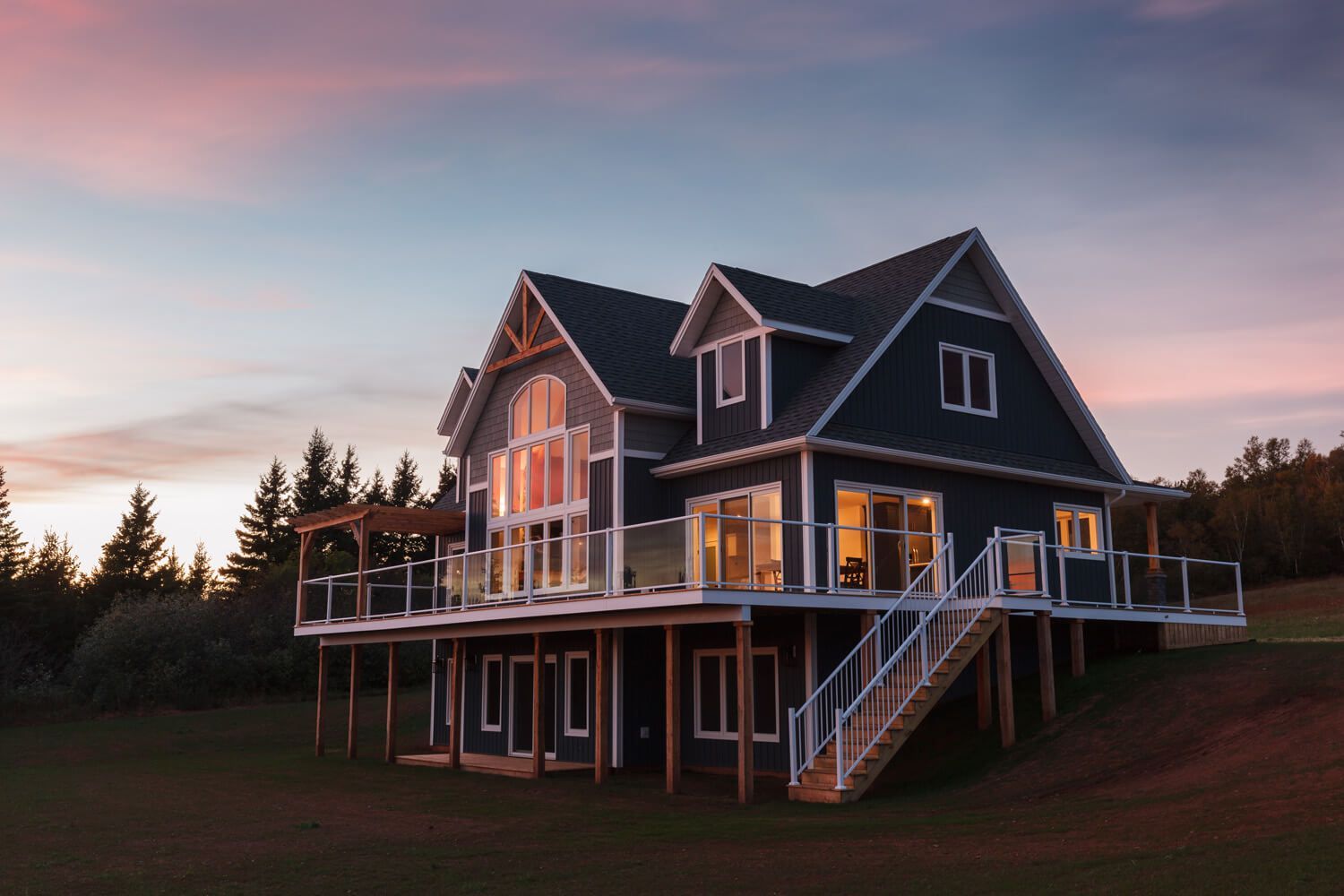 A multi-level dark blue house with a wrap-around deck sits on a grassy hill during a pink and orange sunset.