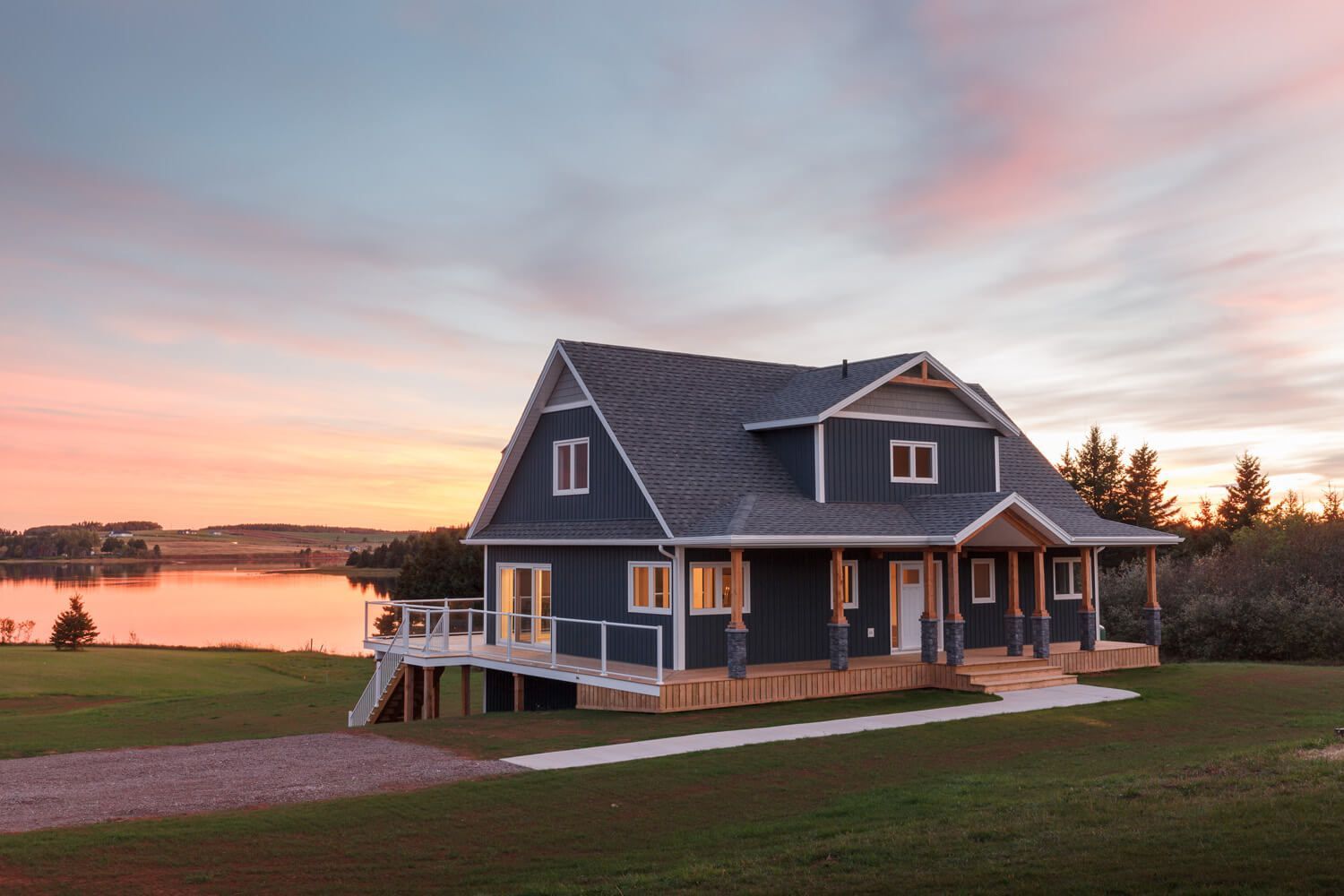 A dark blue two-story house with a wooden porch and deck, situated by a lake at sunset under a soft pink and blue sky.
