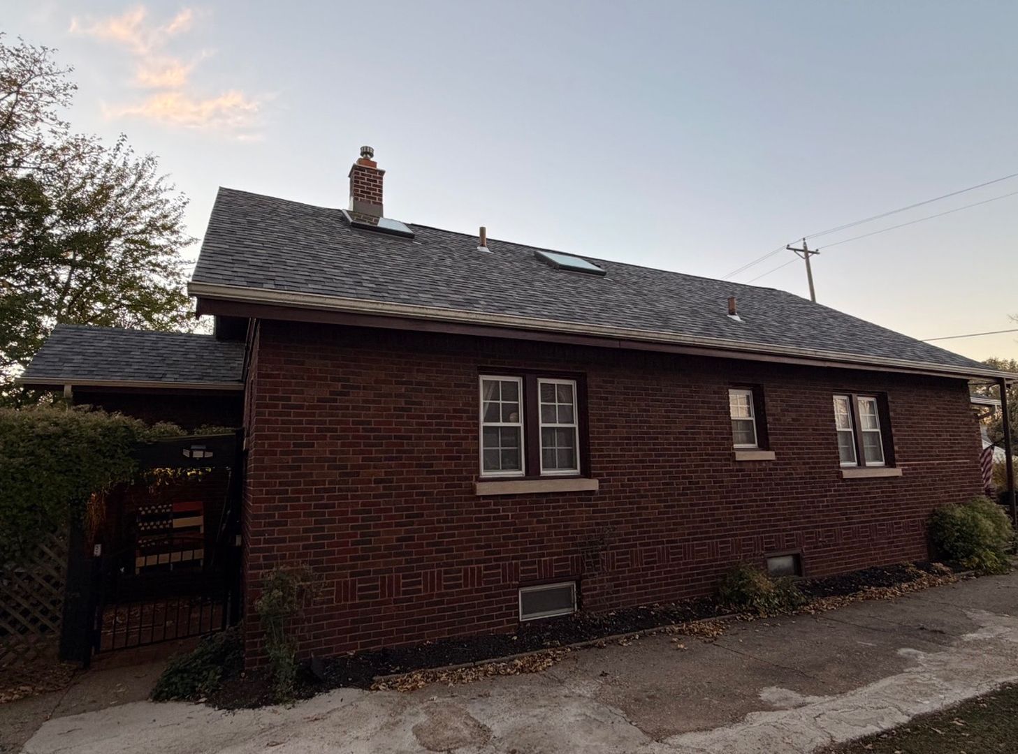 A red brick house with a dark shingled roof, a chimney, and three windows under a pale sky.