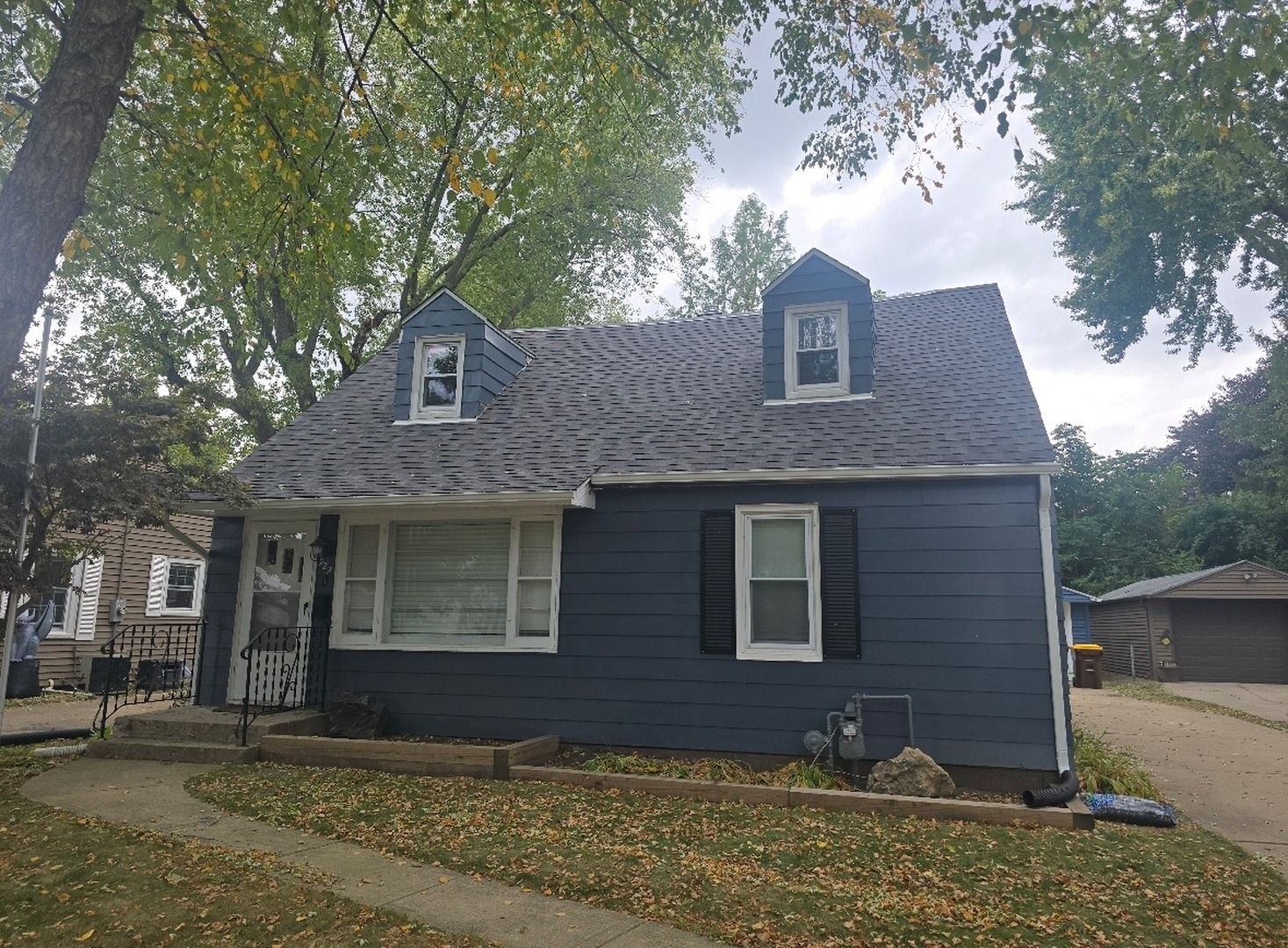 A blue, one-and-a-half-story house with white trim, two roof dormers, and a driveway, surrounded by trees on a cloudy day.