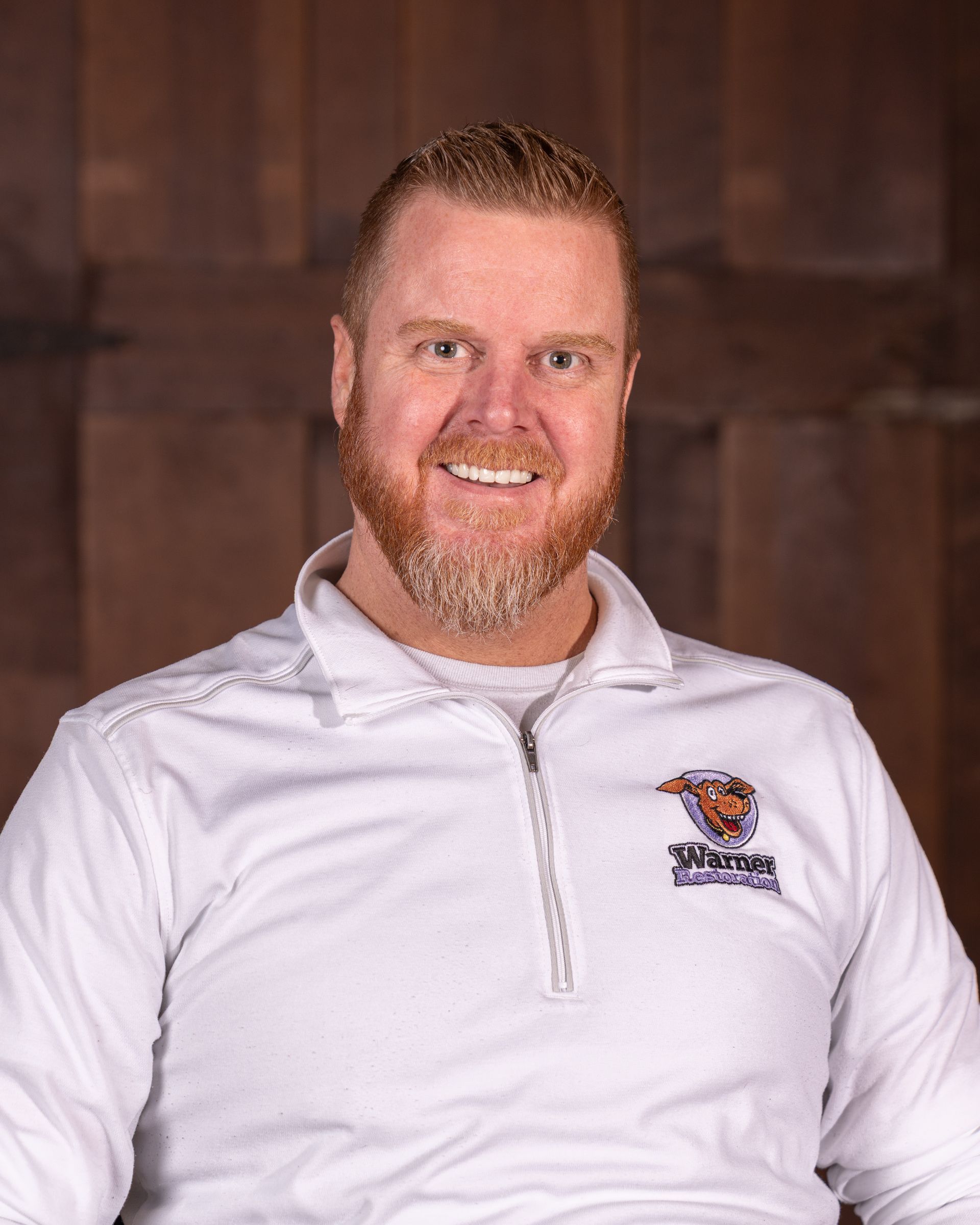 A person with a beard and short hair wearing a white quarter-zip pullover with a logo against a brown wooden background.