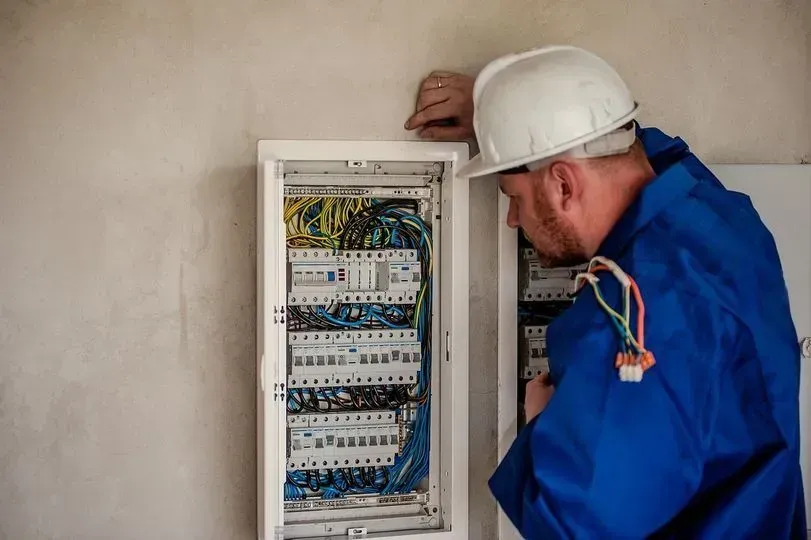 Electrician in blue workwear inspecting an open wall-mounted circuit breaker panel