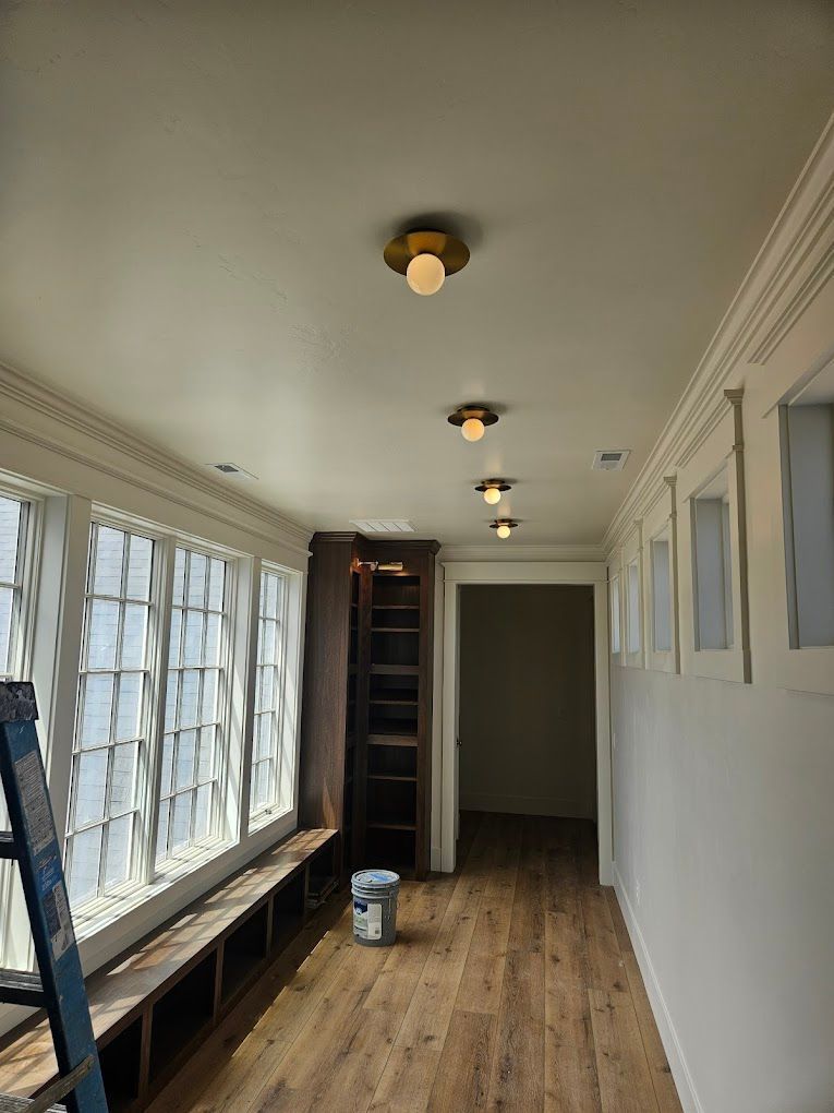 Narrow empty hallway with wood floors, windows on left, white walls, and ceiling lights.