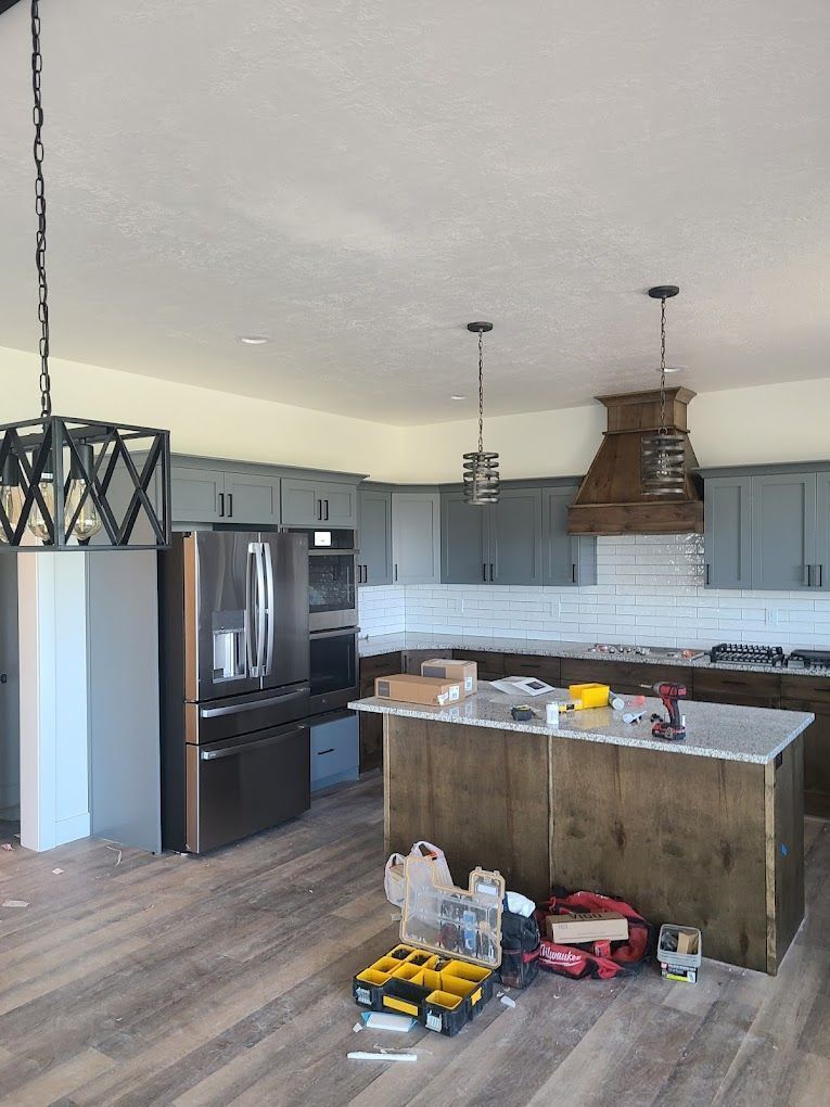 Modern kitchen with dark wood island, stainless fridge, gray cabinets, and pendant lights; boxes and tools on floor.
