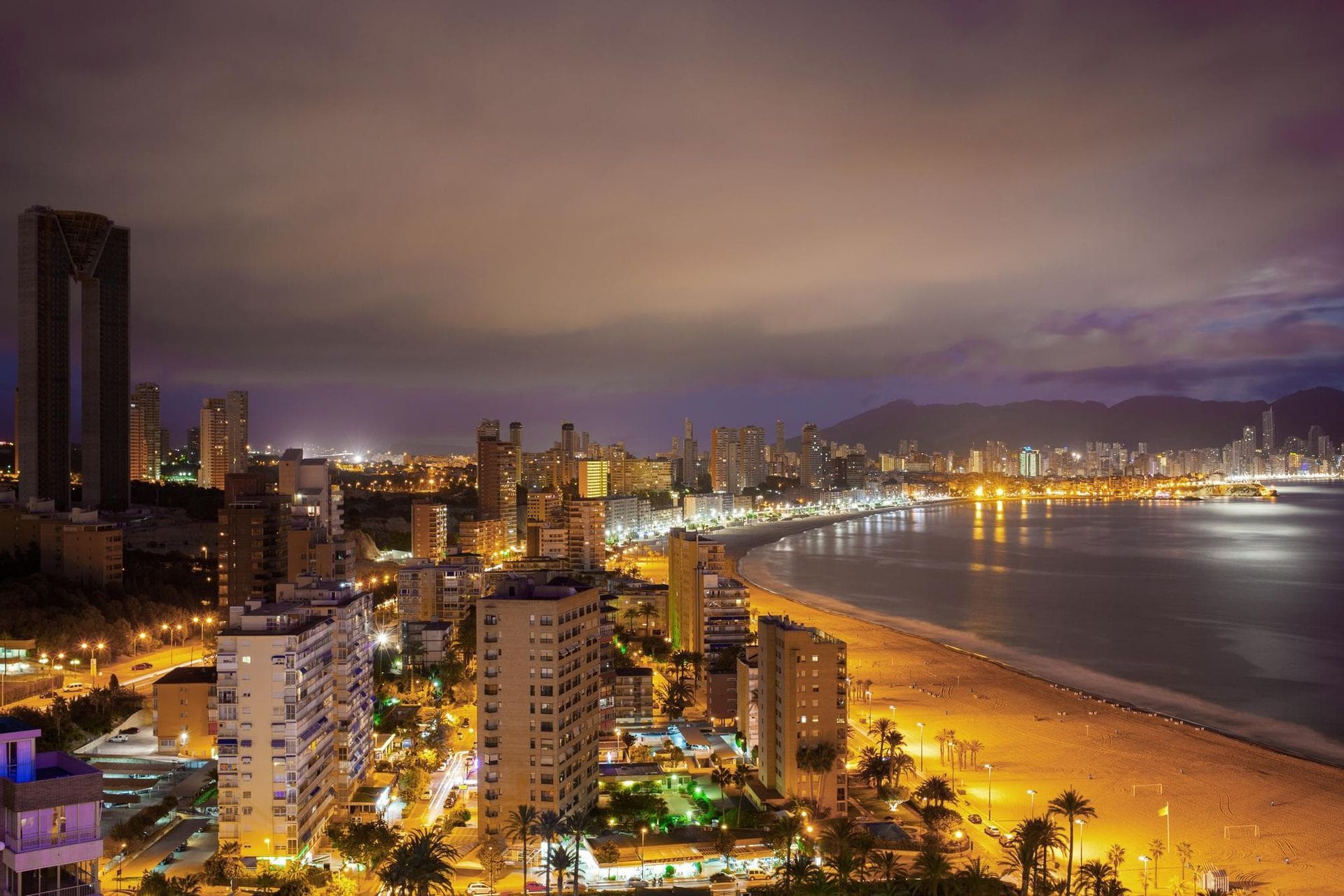 Vista noturna de uma cidade costeira com prédios iluminados, praia e águas calmas sob um céu nublado.