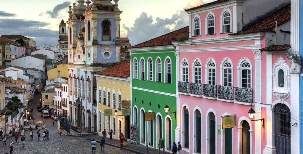 Rua colonial colorida com edifícios na encosta, uma torre de igreja e pessoas caminhando abaixo.