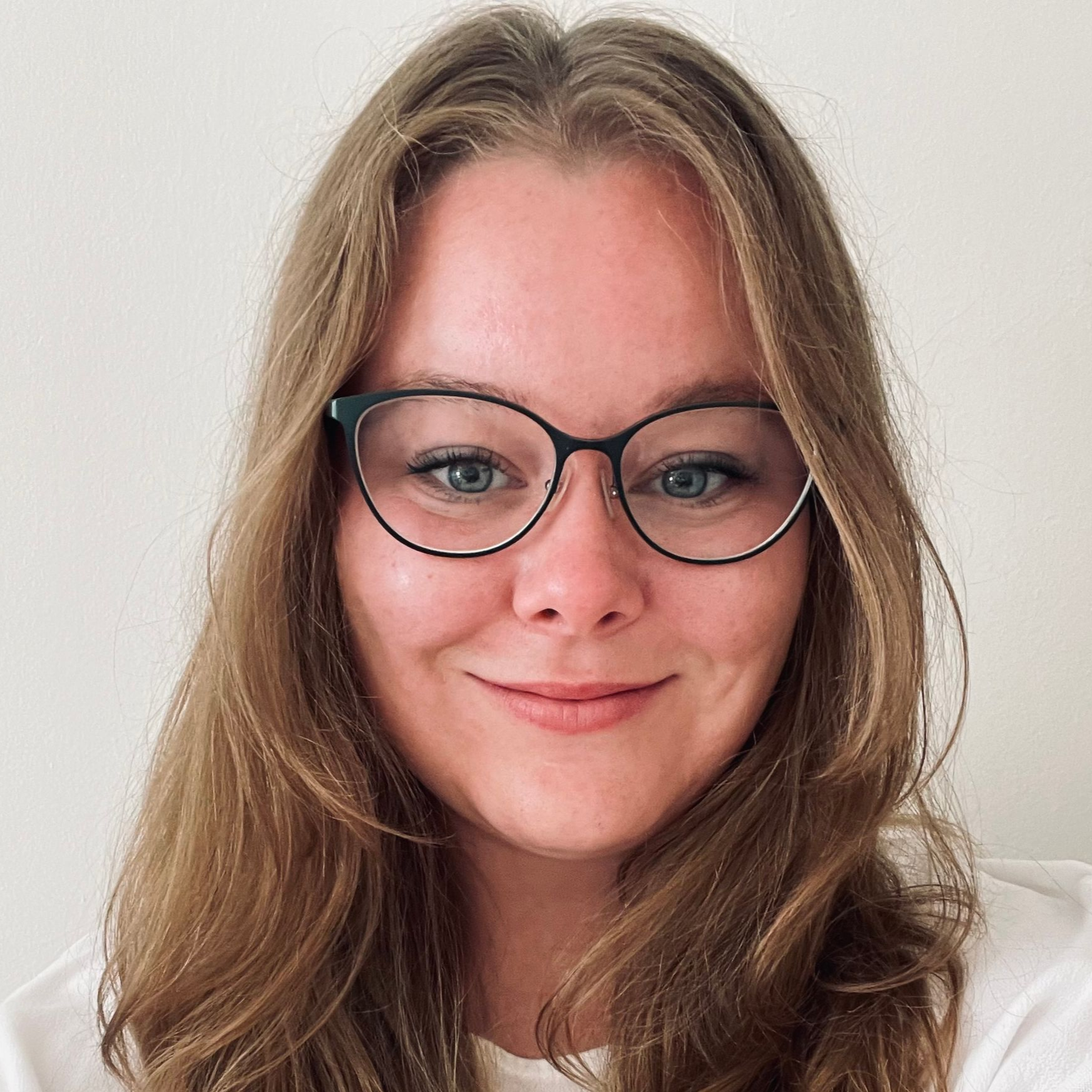 Foto: Esther Heinz, Sie steht vor einem hellen Hintergründ und trägt ein dunkelgraues loose Fit T-Shirt. Esther trägt eine Brille und offene Haare