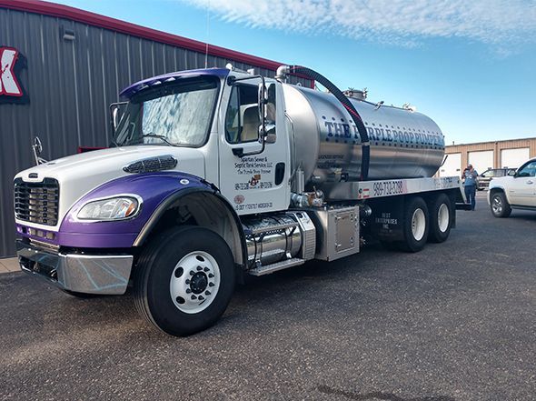 A white and purple vacuum tanker truck parked on an asphalt lot beside a building.