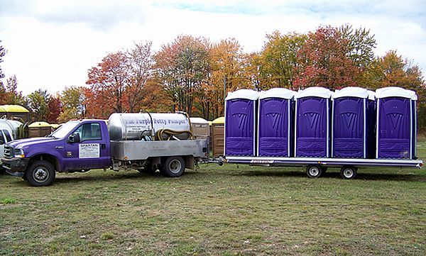 A purple truck with a utility tank pulls a flatbed trailer loaded with five purple portable toilets in a grassy field.