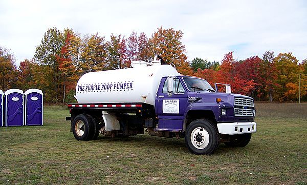 A purple septic pump truck parked in a grassy field next to several blue portable toilets, with colorful autumn trees.