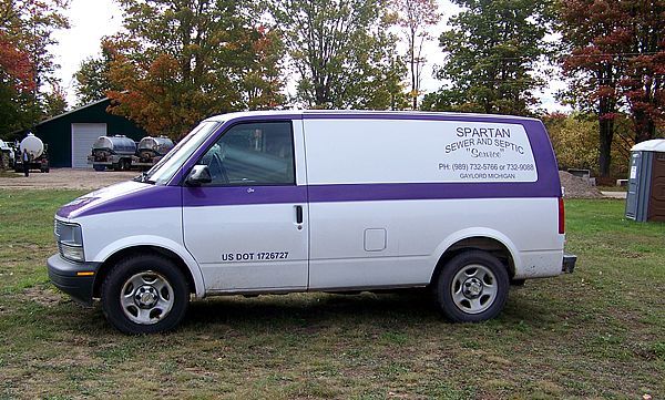 A white and purple utility van parked on grass in a rural setting with trees and a shed in the background.
