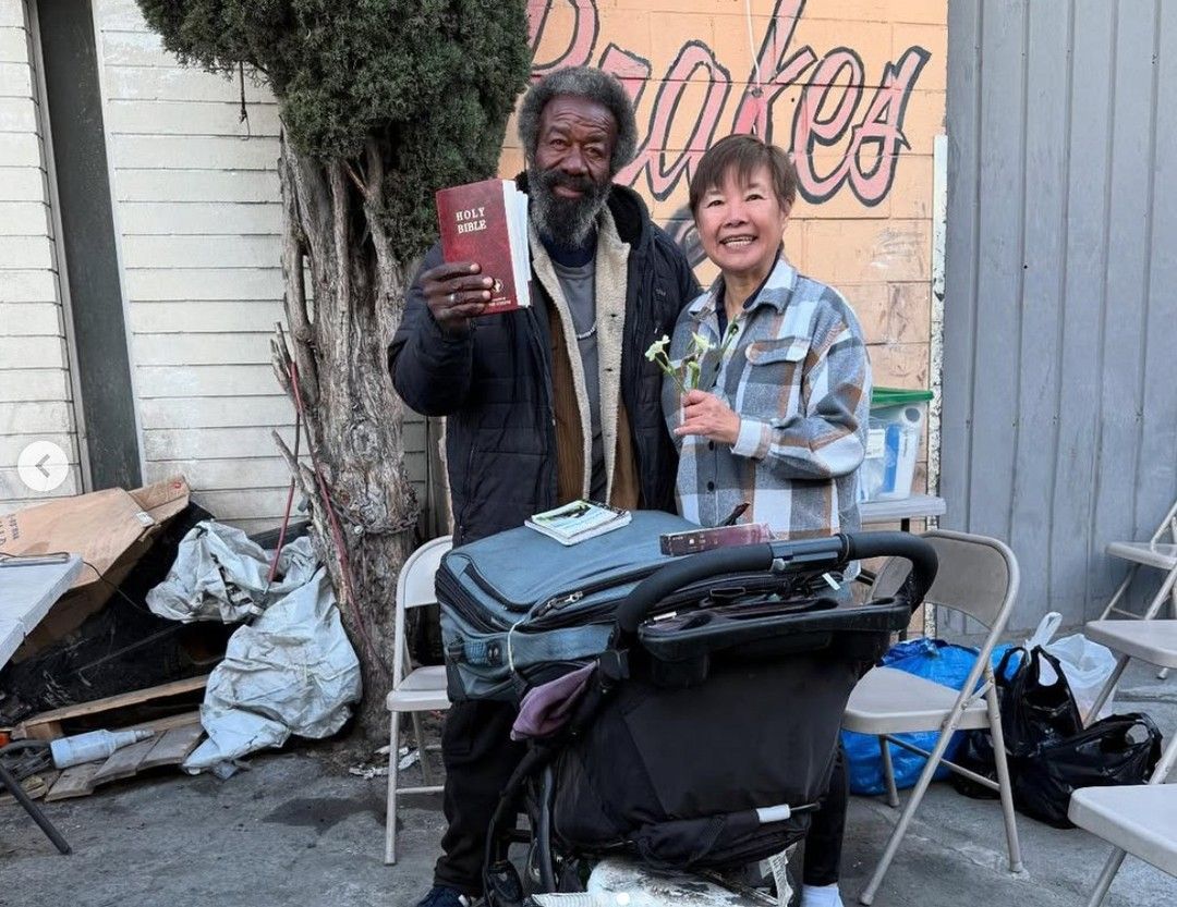 A person with a beard holds up a Bible next to a smiling person holding a flower, standing behind a stroller outdoors.