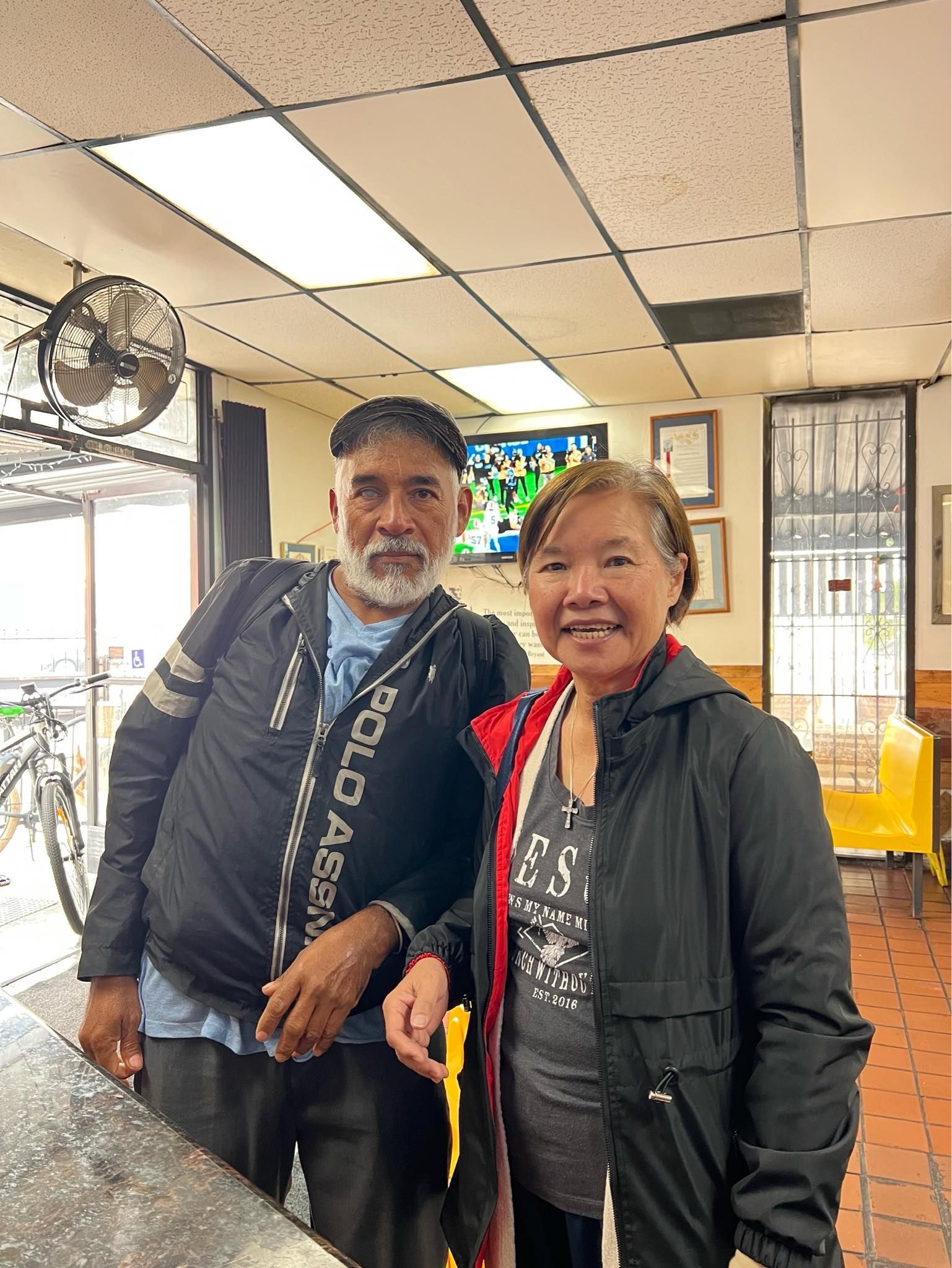 A man with a white beard and a woman stand together inside a shop, smiling for a photo under indoor lighting.