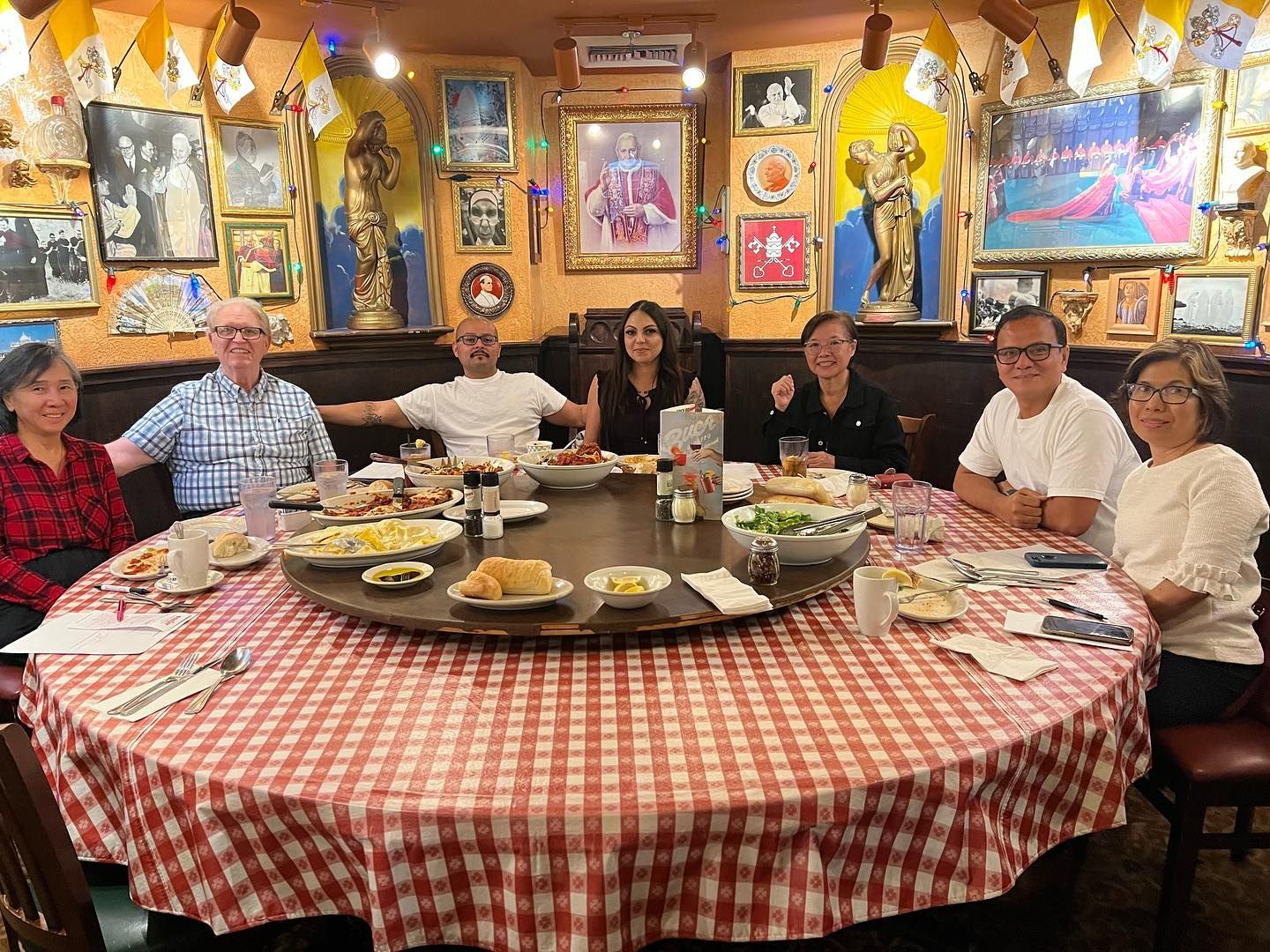 Seven people sit around a large, round table with a red-and-white checkered cloth in a warmly lit, decorated restaurant.