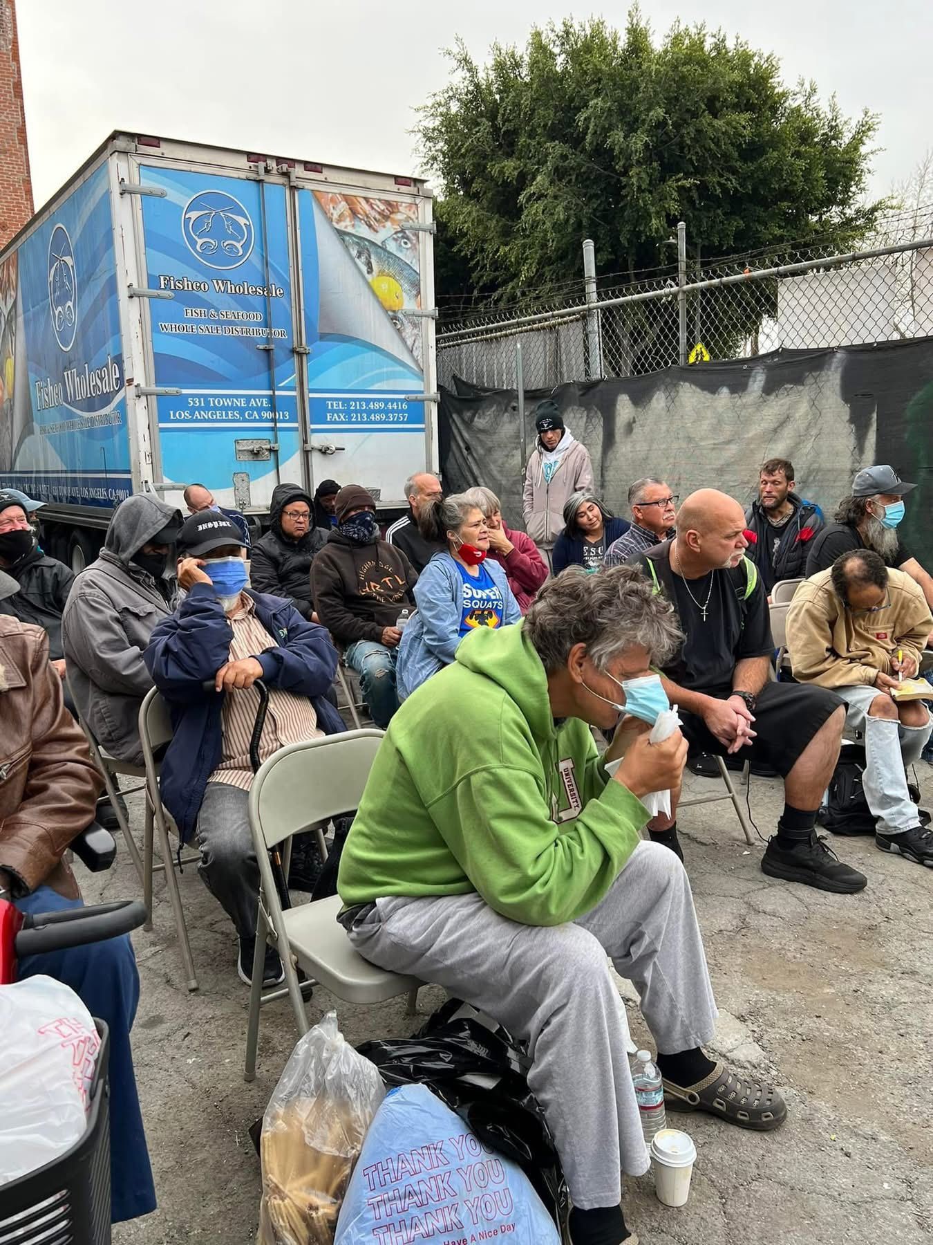 A group of people sit on folding chairs in an outdoor area near a blue semi-truck trailer, some wearing face masks.