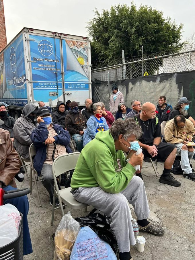 A group of people sit on folding chairs in an outdoor area near a blue semi-truck trailer, with some wearing face masks.