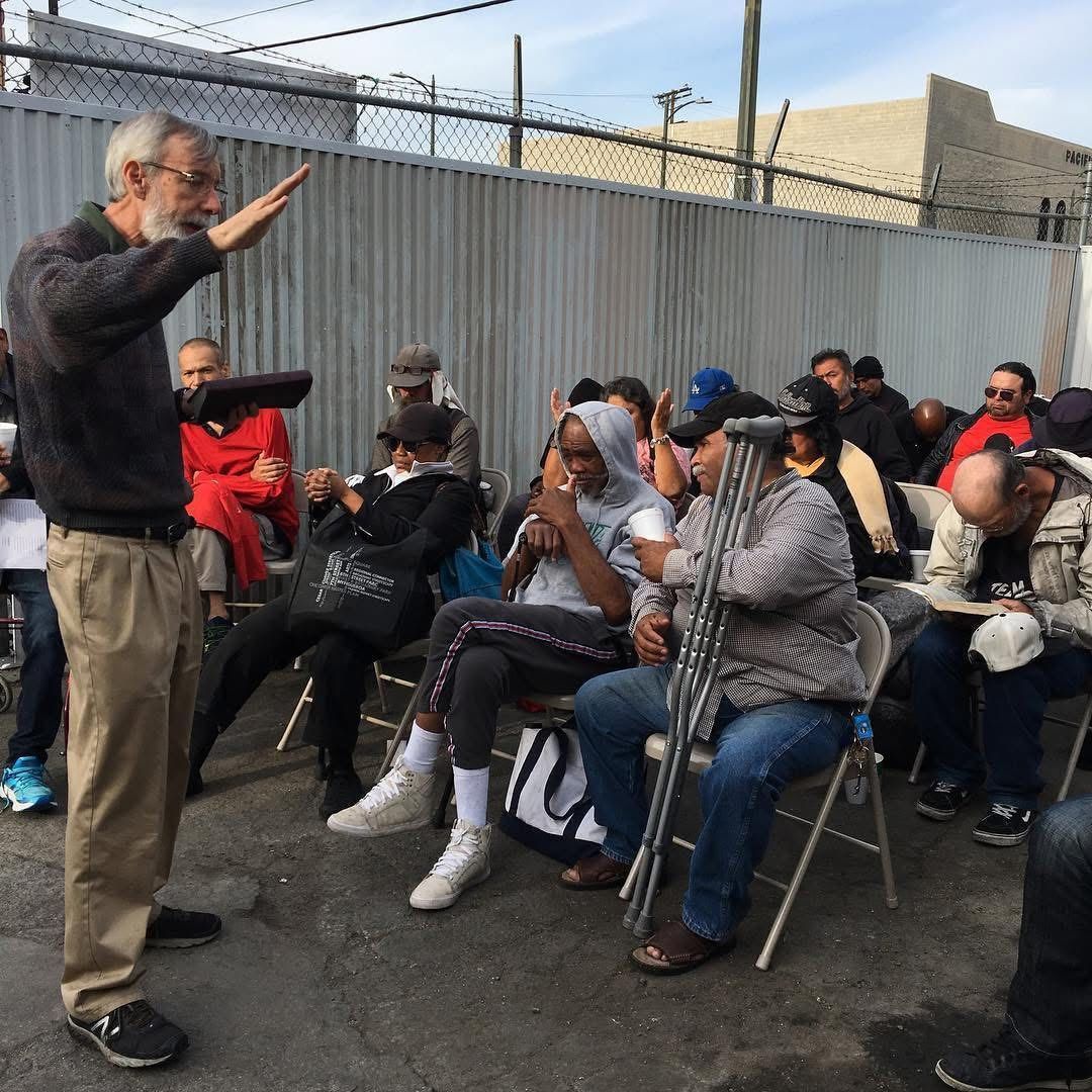 A speaker stands before a seated group outdoors against a chain-link fence, gesturing as he addresses the audience.