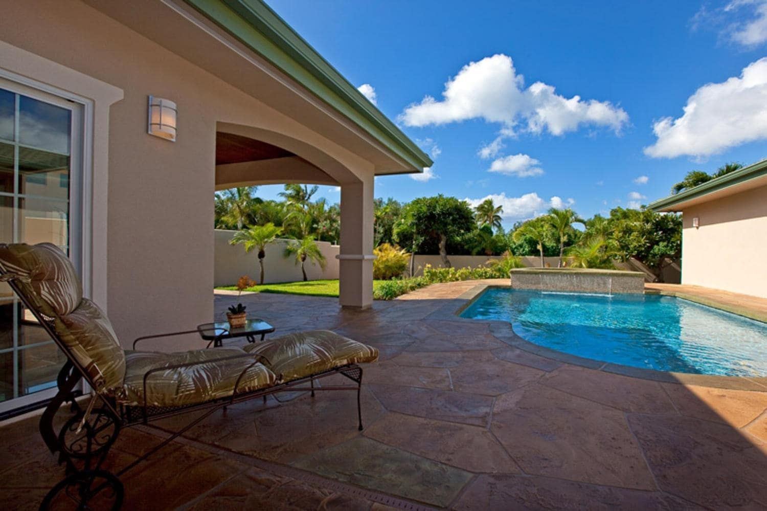 Lounge chair on patio overlooking pool and lawn, with blue sky and clouds.