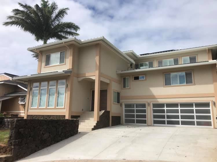 Beige two-story house with glass garage doors, windows, and palm tree on the roof, with a concrete driveway and cloudy sky.