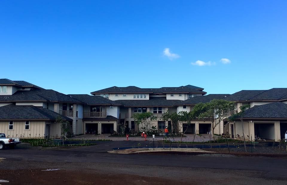 Multi-story beige and dark roofed building with a blue sky. A dark paved area is in front.