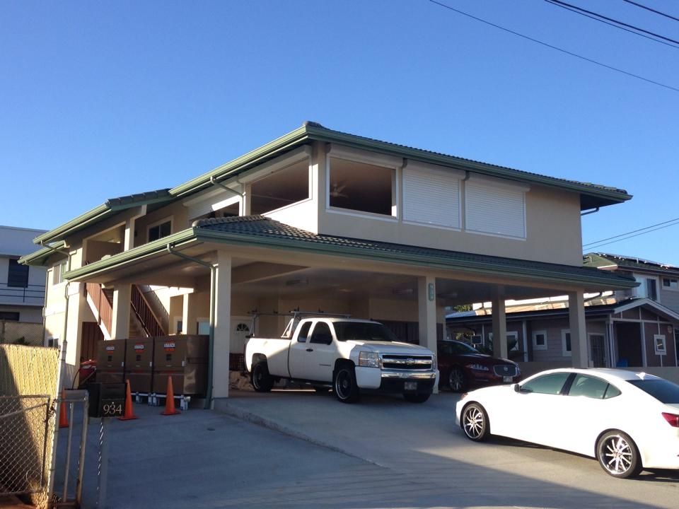 Two-story building with covered parking. White pickup truck and car parked under. Green roof, blue sky.