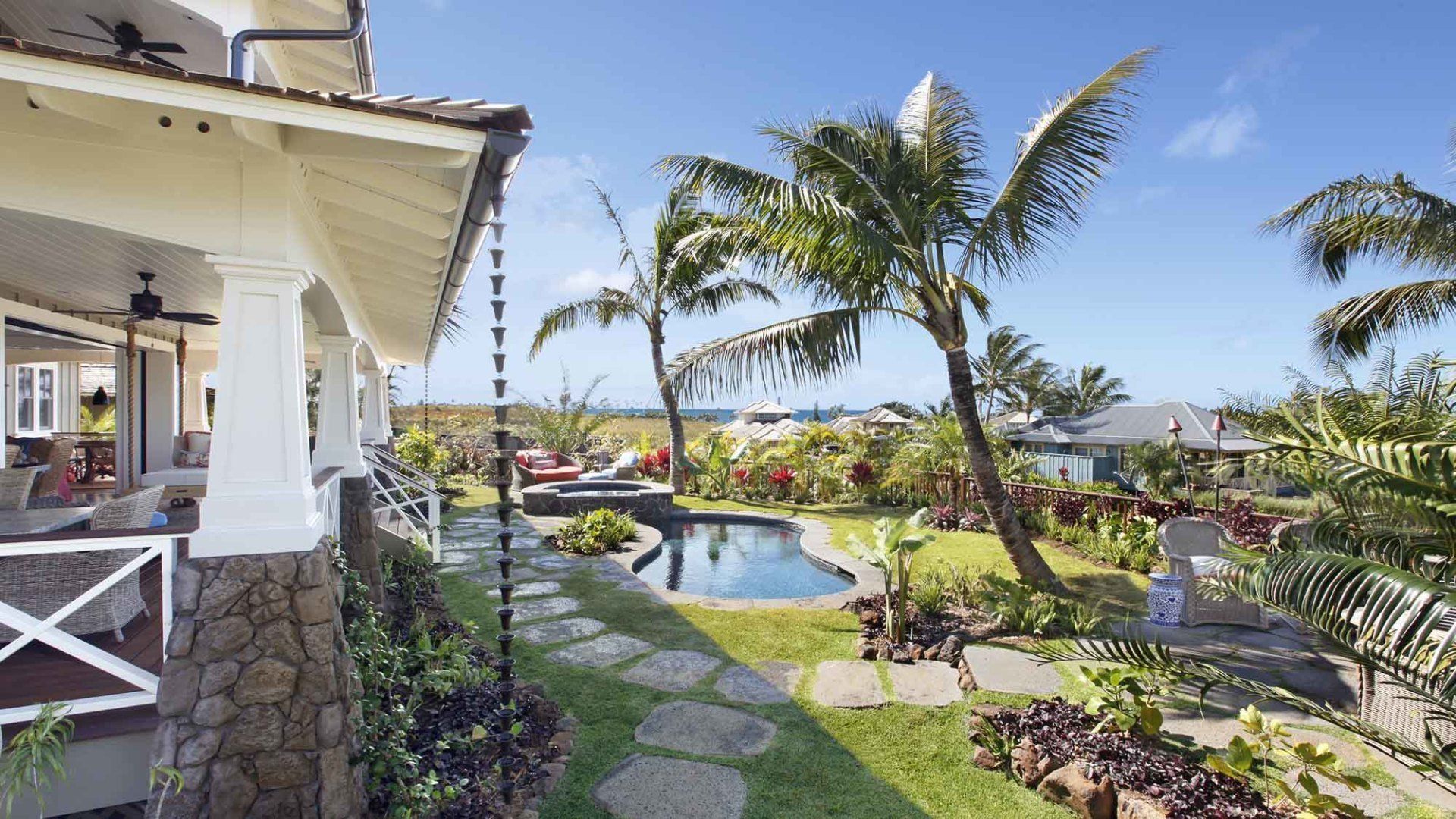 Tropical house exterior with a porch overlooking a pool, palm trees, and lush landscaping. Blue sky.