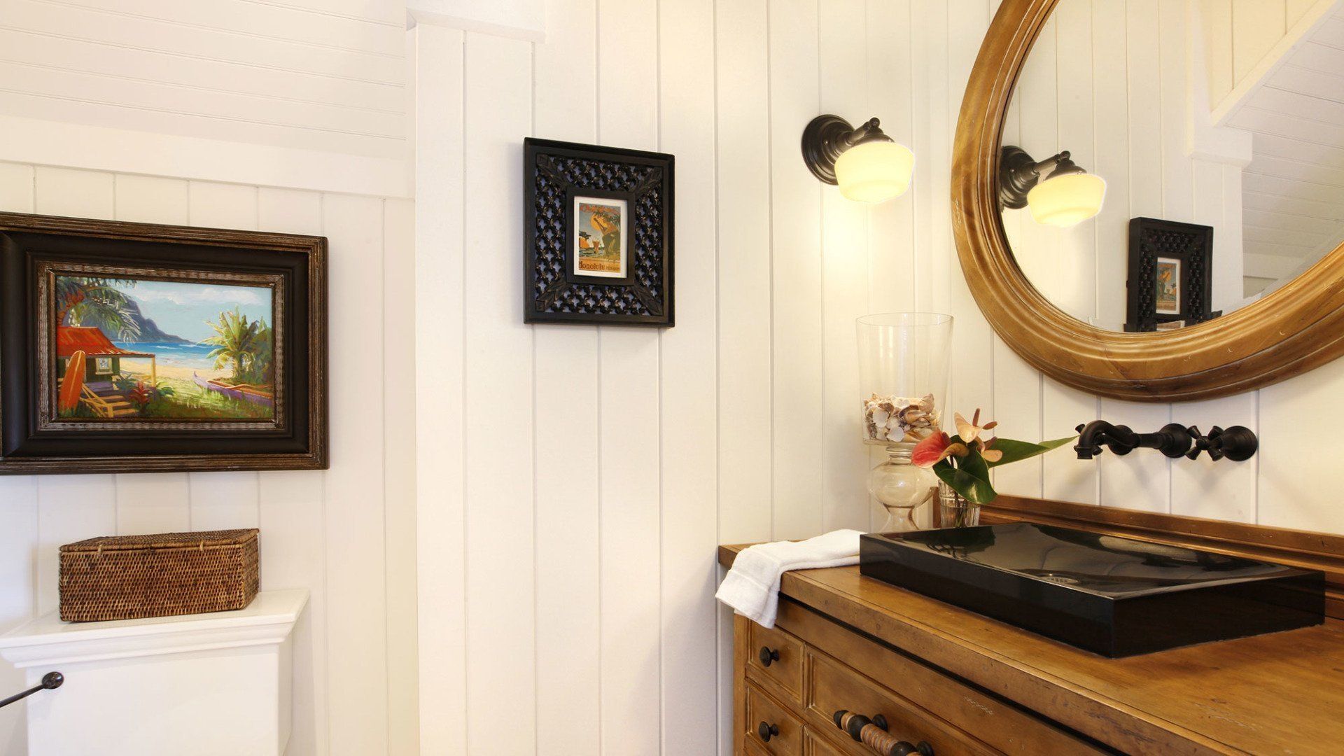 A bathroom with a wooden vanity, a black sink, a round mirror, and framed art. White shiplap walls.
