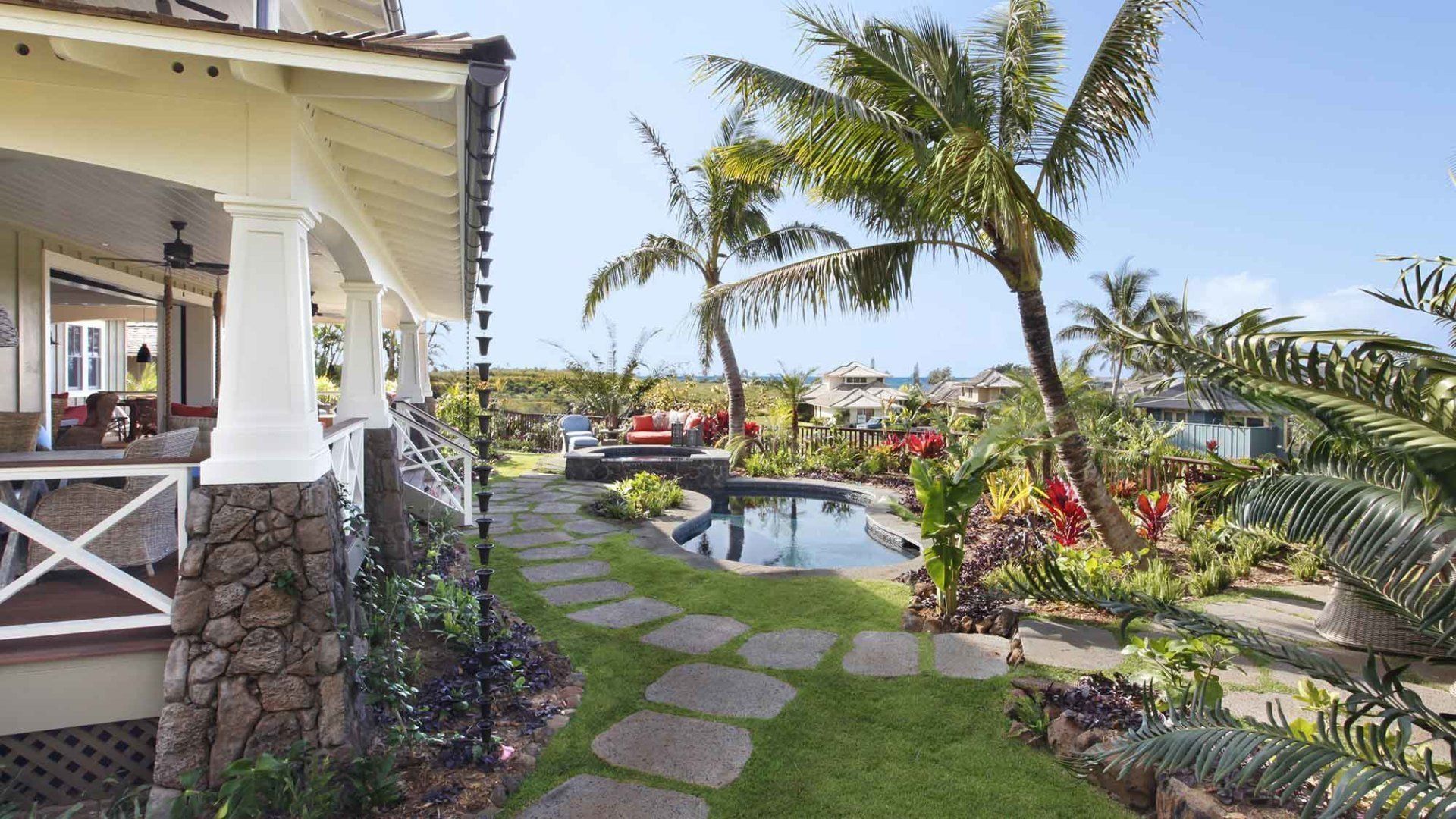 Stone path leads to pool and tropical garden, next to a house with a covered porch.