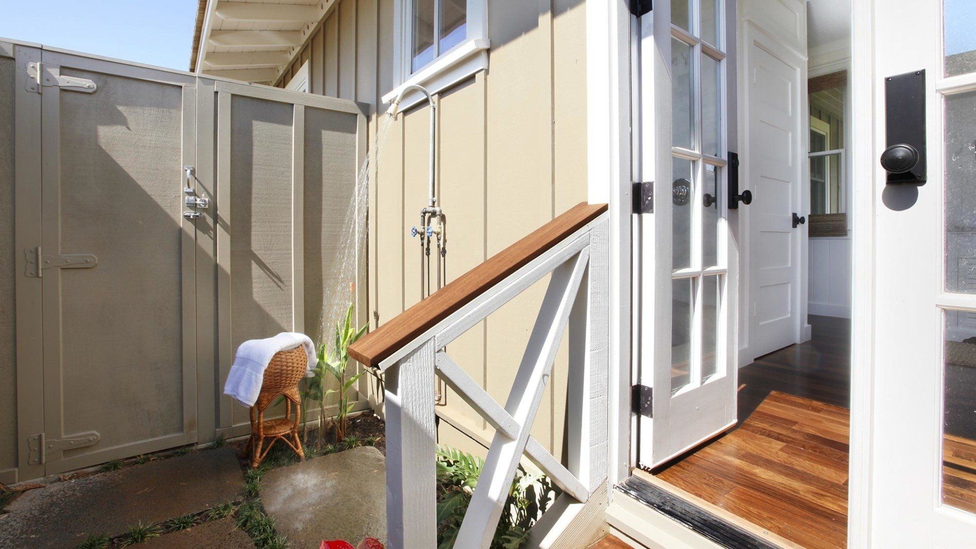 Outdoor shower with beige siding, wooden deck, white doors, and a small chair with a towel.