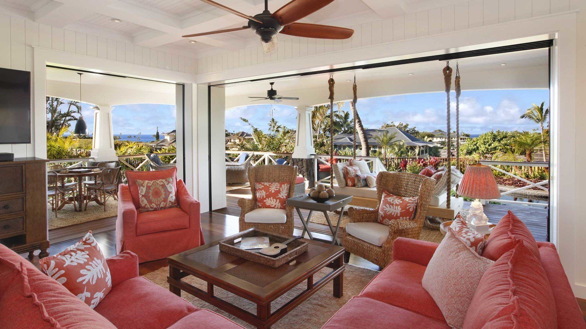 Living room opens to a balcony with ocean view, red couches and wicker chairs, wood ceiling fan.
