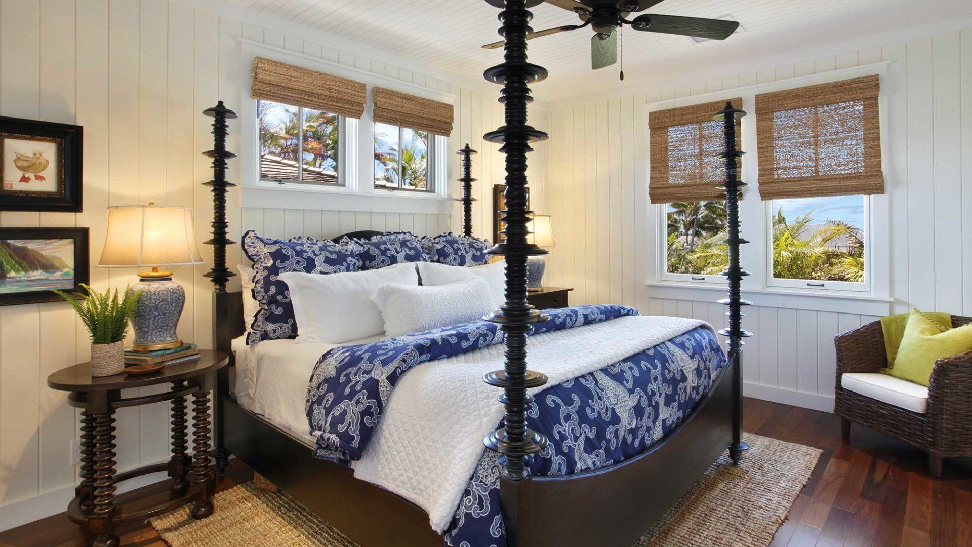 Bedroom with four-poster bed, blue and white bedding, windows with blinds, and side table with lamp.