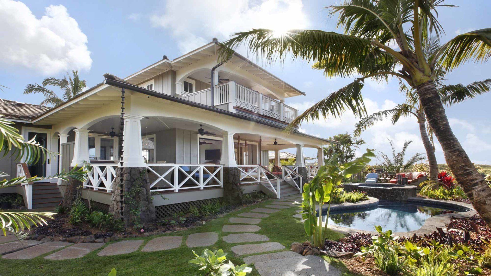 Tropical two-story house with porch, balcony, and pool, stone pathway, surrounded by palm trees and lush landscaping.