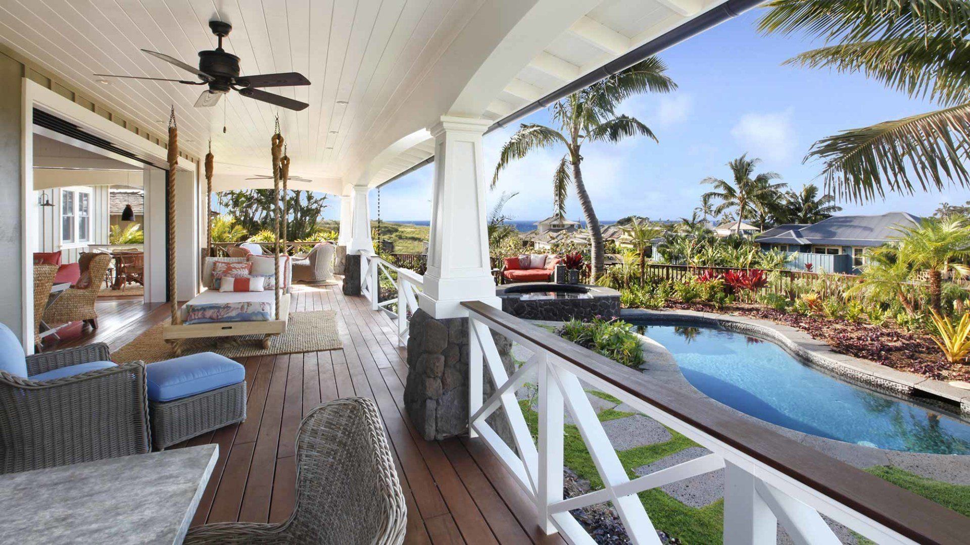 Wooden porch overlooking pool and tropical landscape. Blue sky, palm trees, outdoor seating.
