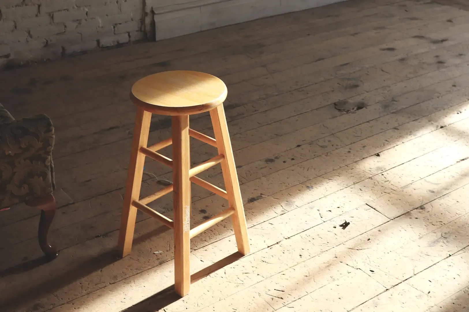 A solitary wooden stool standing on a rustic, sunlit wooden floor against a weathered brick and plas