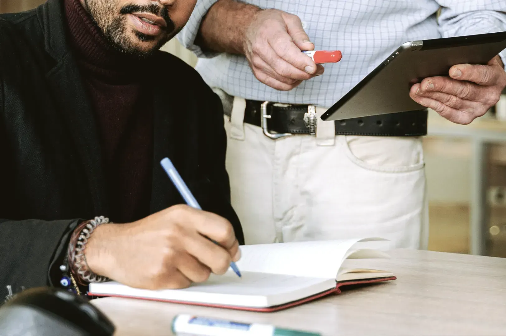 A mentor holding a digital tablet while coaching a professional who is taking notes in a notebook, i