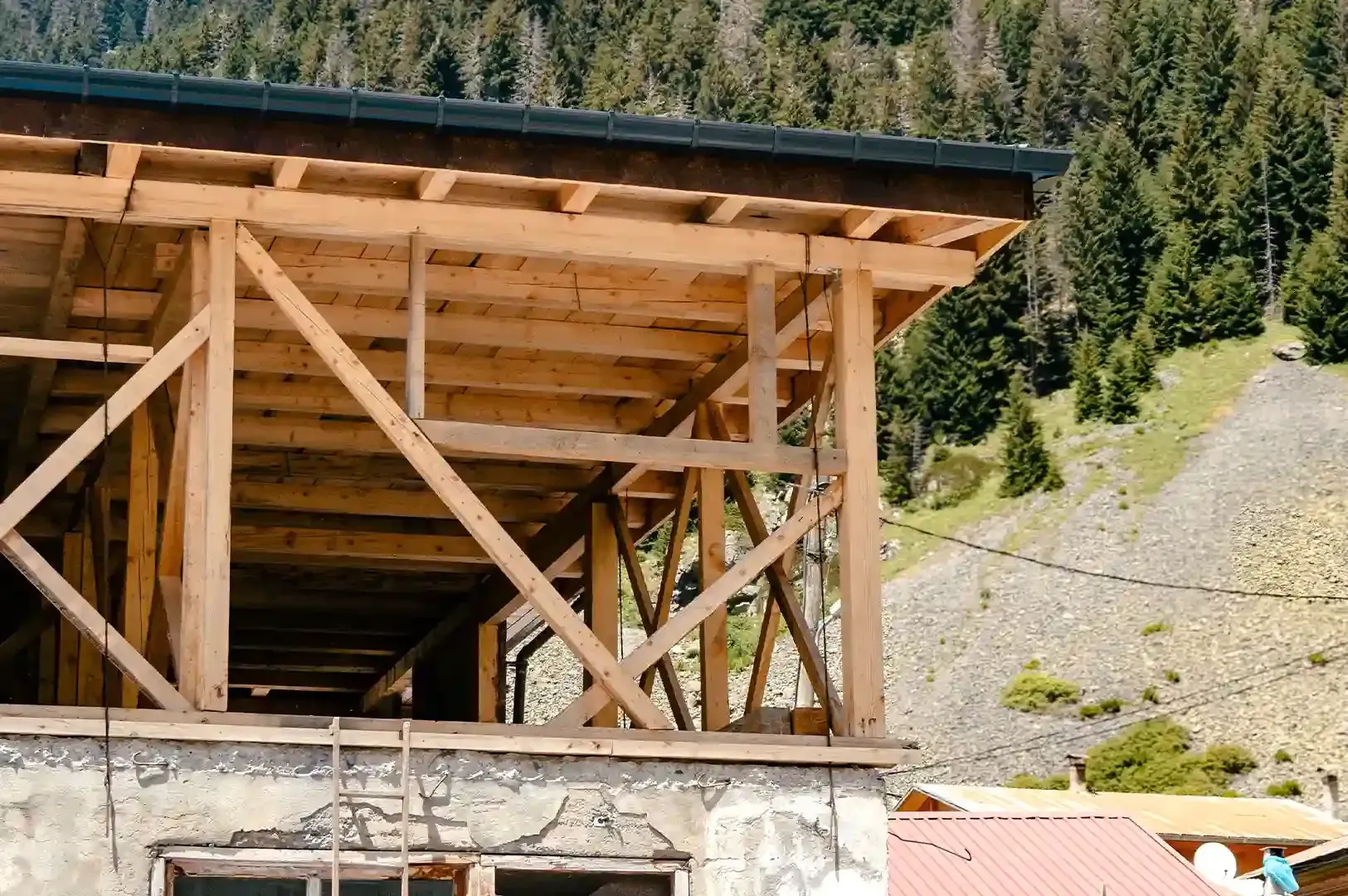 An exterior view of an exposed timber-framed roof structure under construction against a mountain an