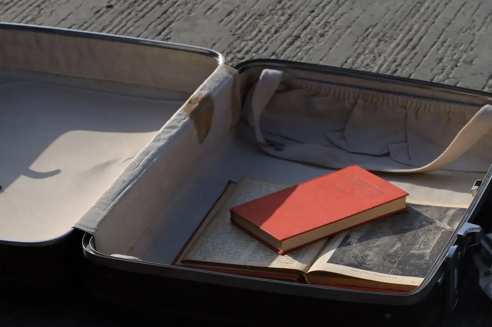 An open vintage suitcase resting on a textured surface, containing only an open book and a bright re